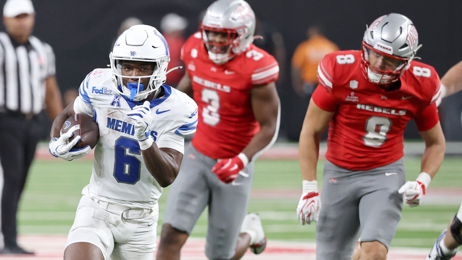 A football player in a white and blue Memphis jersey (#6) carries the ball while being pursued by two players in red Rebels jerseys during a game.