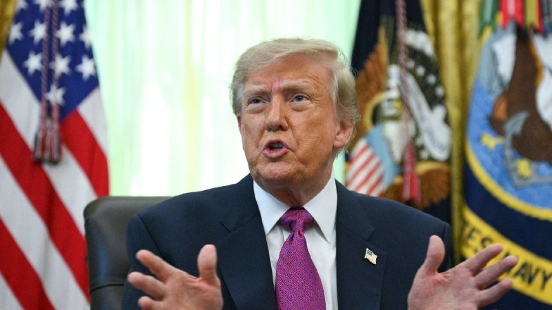 A blonde-haired man in a dark suit and pink patterned tie speaks with raised hands in front of an American flag and official emblems.