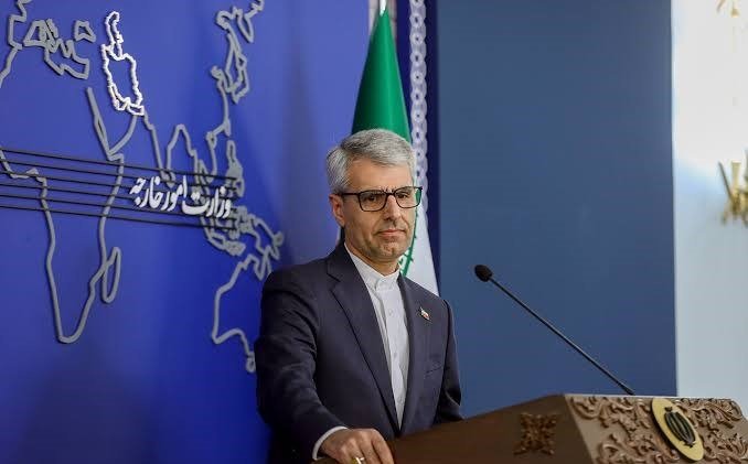 A gray-haired man in a dark suit stands at a carved wooden podium with a microphone, speaking before a blue backdrop featuring a world map outline.