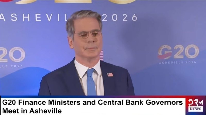 A gray-haired man in a blue suit and glasses speaks at a podium against a blue "G20 Asheville 2026" backdrop.