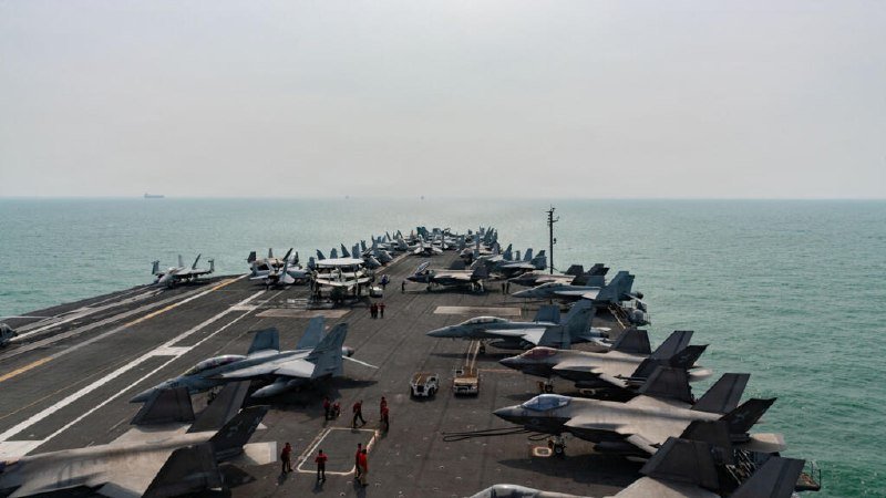 Military fighter jets are parked in rows on an aircraft carrier's flight deck at sea, with crew members in colored vests standing among the aircraft under a hazy sky.