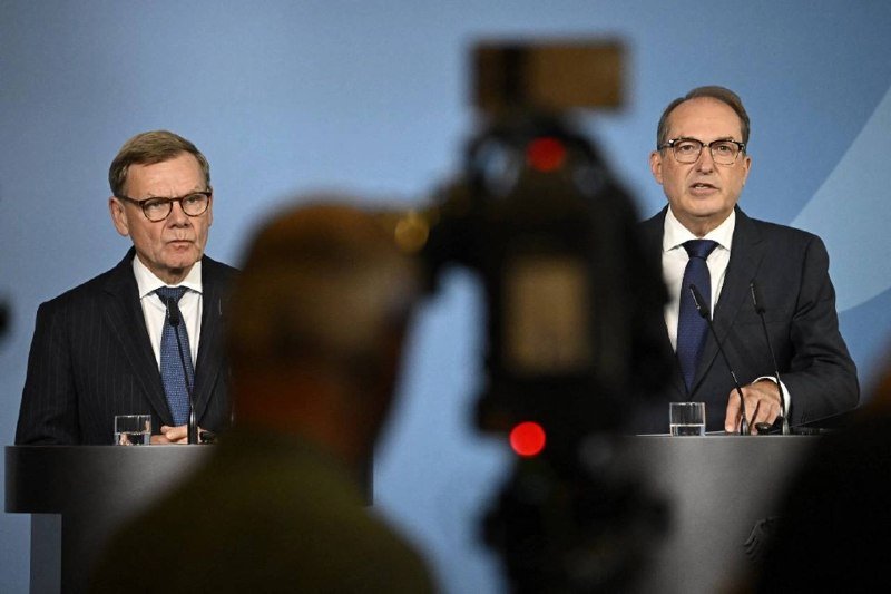 Two men in dark suits stand at podiums against a blue backdrop, with one speaking into a microphone while a camera films from the foreground.