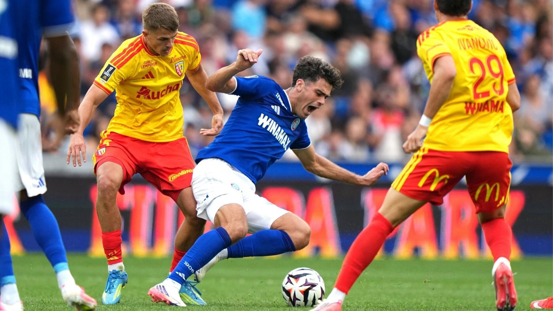 A soccer player in a blue jersey falls while being challenged by two opponents in yellow and red uniforms during a match.