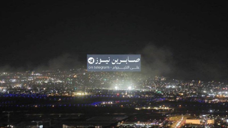 A nighttime aerial view shows a sprawling city illuminated with lights and smoke plumes rising in the background, overlaid with an Arabic Telegram logo and text.