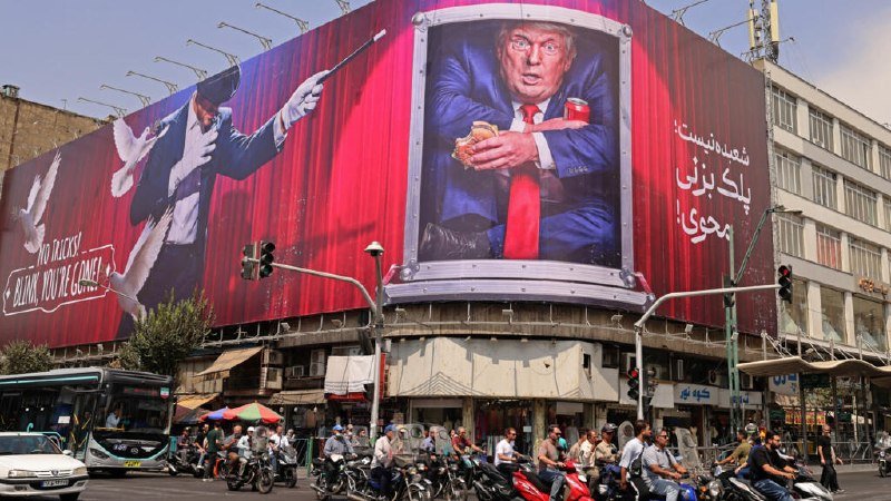 A large red billboard displaying a caricature of a man in a suit eating a sandwich and holding a coffee cup is mounted on a building corner above a busy street filled with motorcyclists and pedestrians.