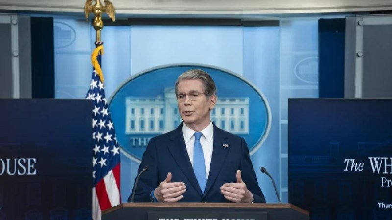 A man in a dark suit and blue tie speaks at a podium in the White House press briefing room, gesturing with both hands before an American flag and the White House seal.