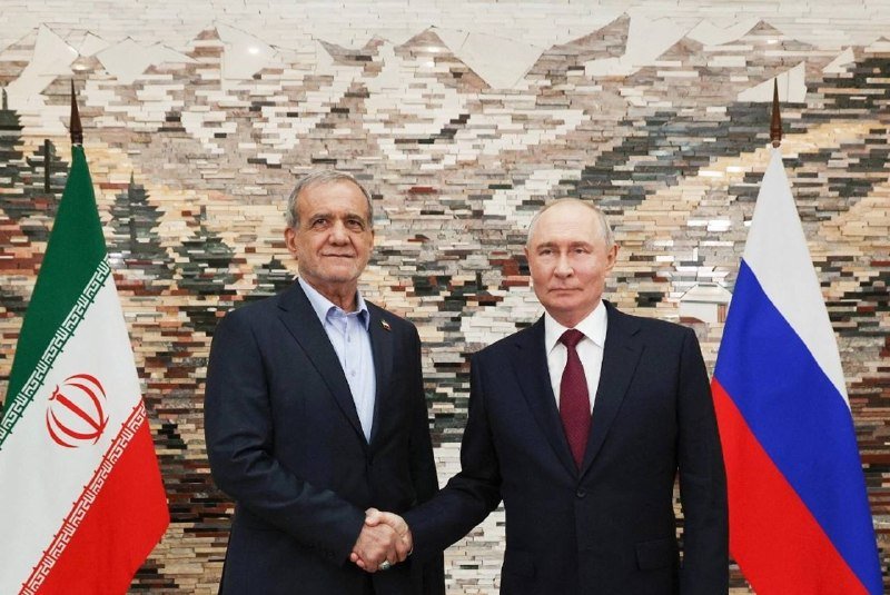 Two men in dark suits shake hands in front of an Iranian flag, a Russian flag, and a textured stone wall.
