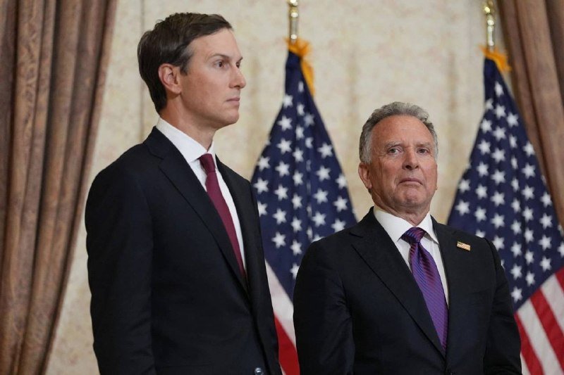 Two men in dark suits stand before two American flags and draped curtains, looking forward with serious expressions.
