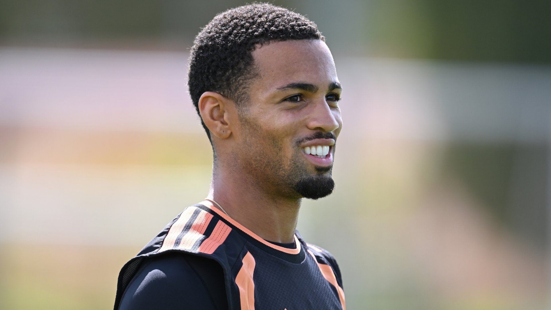 A smiling man with short curly hair and a goatee wears a black and orange athletic top during an outdoor training session.