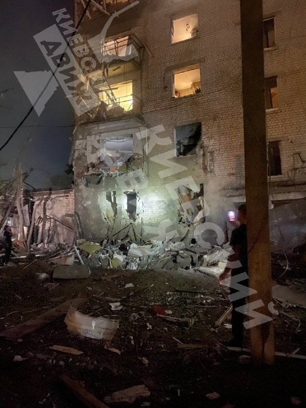 A partially destroyed multi-story brick building stands at night, its lower floors collapsed into rubble with debris scattered around the site.