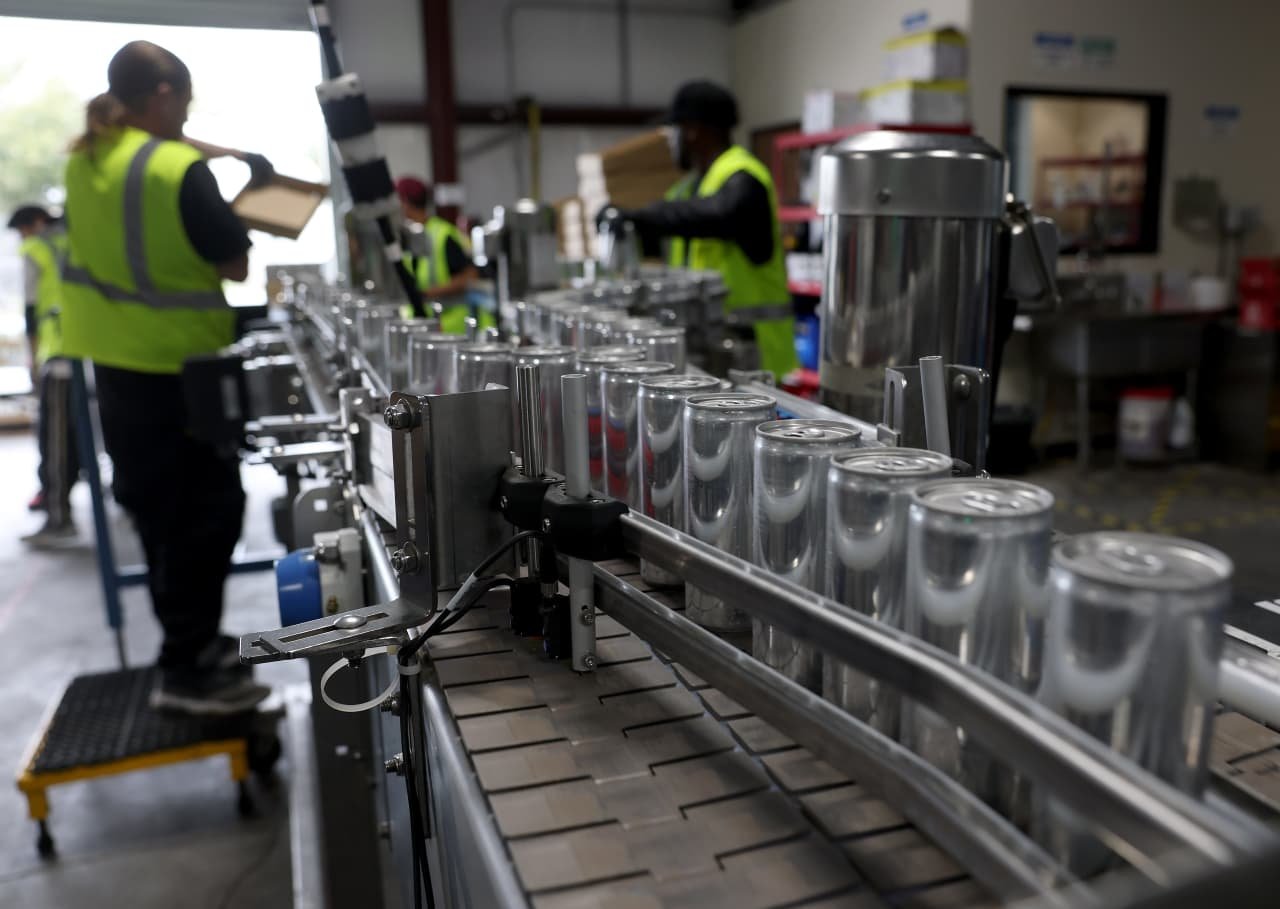Workers in high-visibility vests pack empty aluminum cans onto a conveyor belt in a beverage canning facility.
