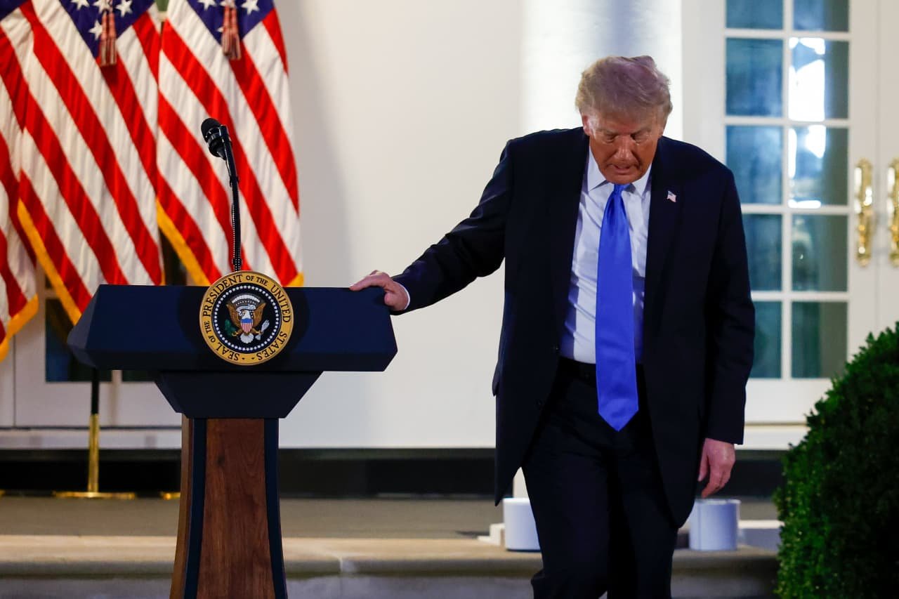 A man in a dark suit and blue tie stands beside a podium bearing the Presidential Seal, with American flags visible behind him.