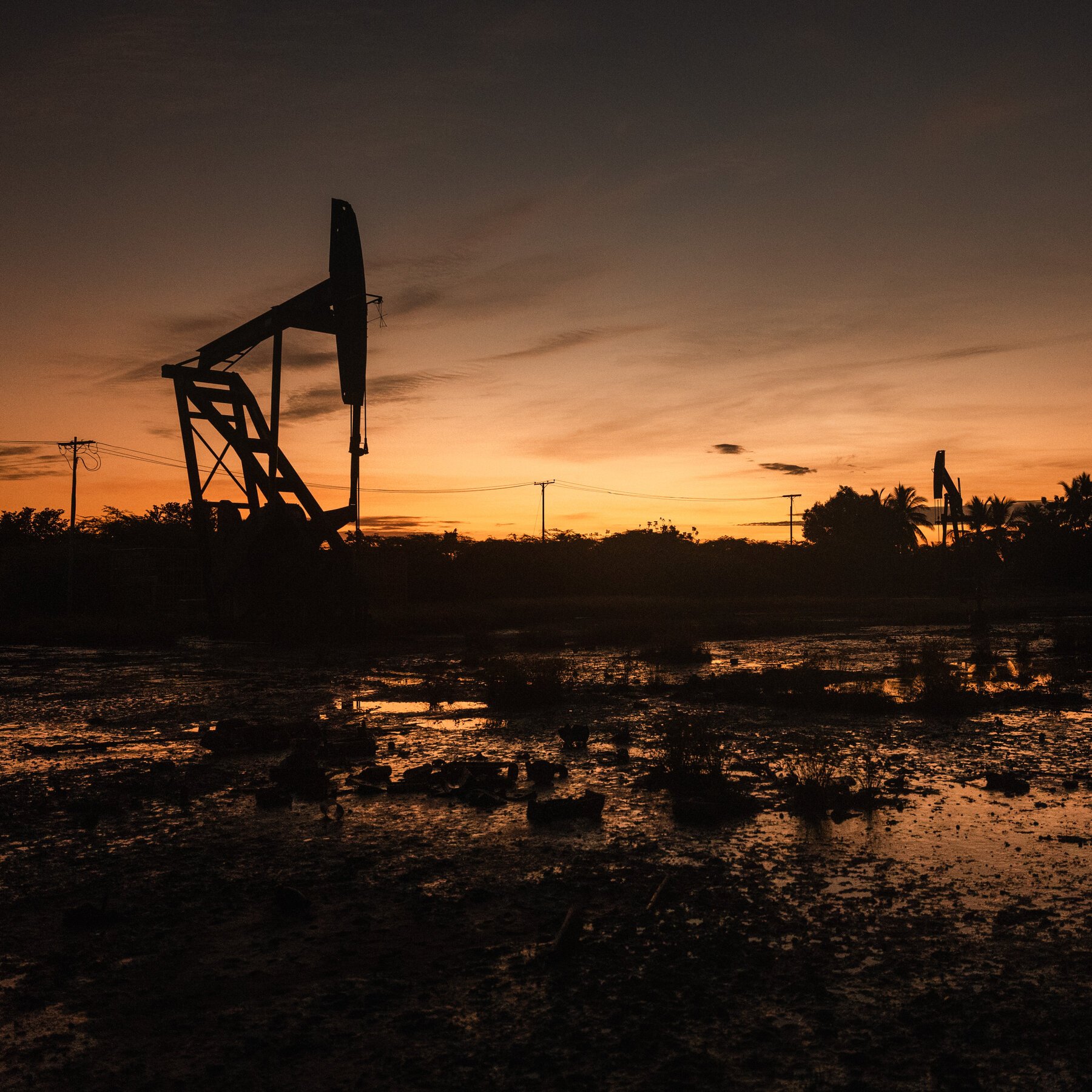 Silhouetted oil pumpjacks stand against an orange sunset sky above a waterlogged, debris-strewn landscape with power lines stretching across the horizon.