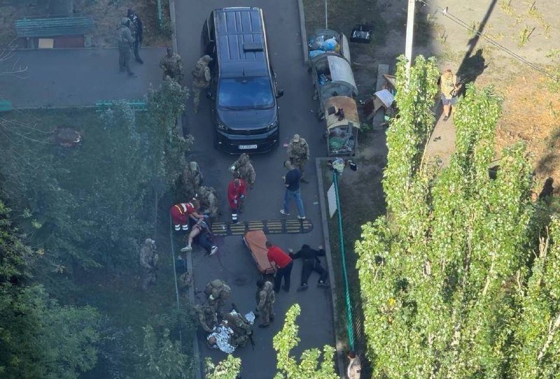 Aerial view shows uniformed personnel, individuals in civilian clothing, and a stretcher on a paved path beside a parked SUV, with scattered debris and greenery along the edges.