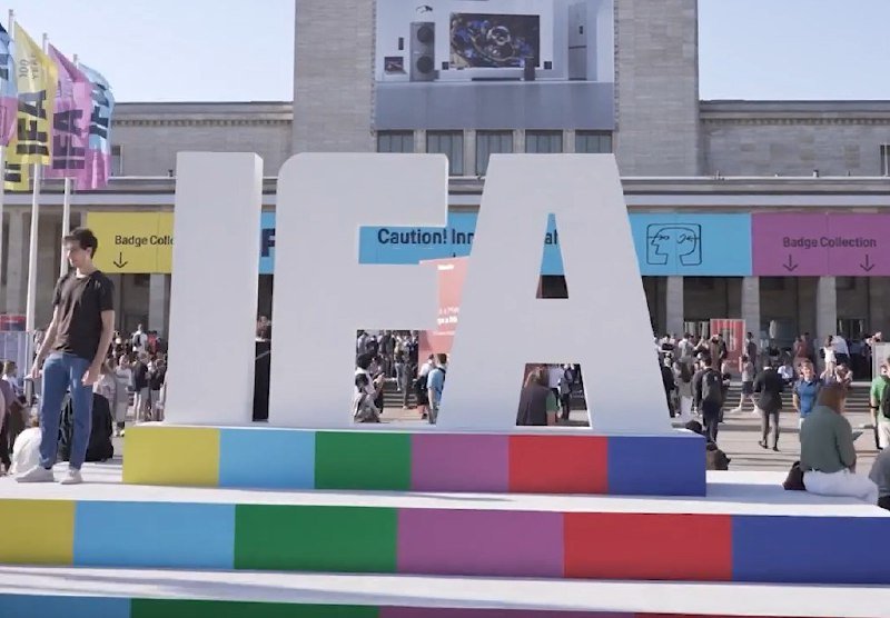 Large white "IAA" letters sit on a multicolored striped platform outside a building, with crowds of people and "Badge Collection" signage visible in the background.
