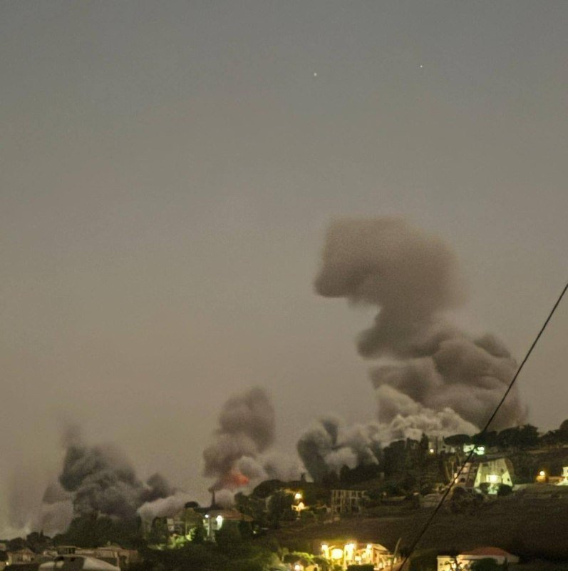 A cityscape at dusk shows multiple plumes of thick gray smoke rising above lit buildings on a hillside.
