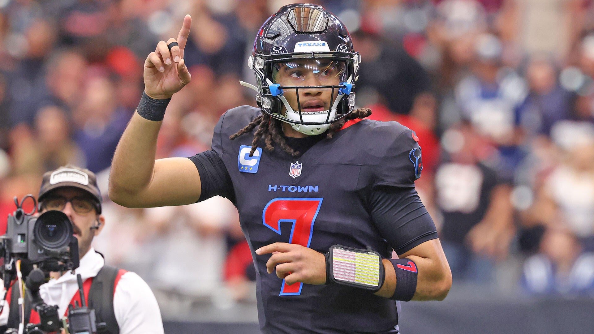 A football player wearing a dark "H-TOWN" jersey with the number 7 and a Texans helmet points skyward on the field, with a blurred crowd and a cameraman visible behind him.