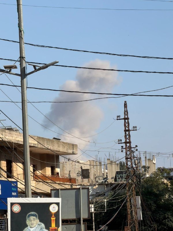A large plume of gray smoke rises above concrete buildings and a tangle of utility wires and a metal tower against a blue sky.
