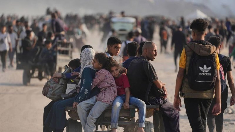 A group of people, including children, ride on a wooden cart through a dusty, crowded outdoor area with many others gathered in the background.