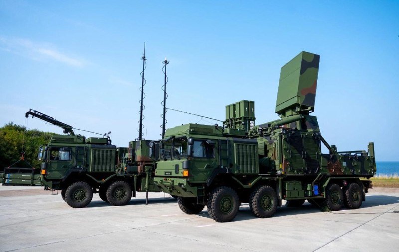 Two camouflaged military radar vehicles with tall antenna masts and a large angled radar panel are parked side by side on a concrete surface near the coast.