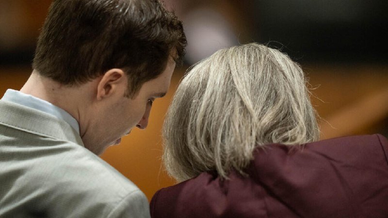 A young man in a light gray suit leans his head toward an older woman with shoulder-length gray hair wearing a maroon jacket.