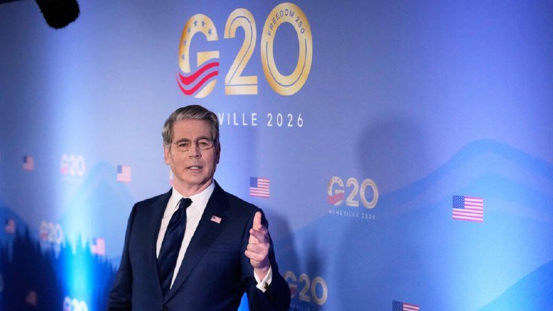 A gray-haired man in a dark suit with an American flag lapel pin speaks and gestures in front of a blue backdrop displaying G20 and American flag logos.