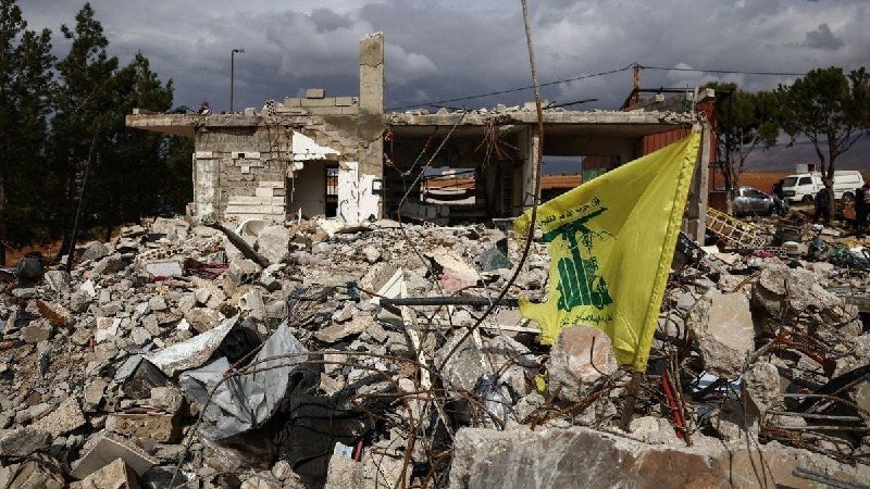 A damaged concrete building with broken walls sits behind a large pile of rubble and debris, with a yellow flag bearing a green emblem planted in the wreckage.