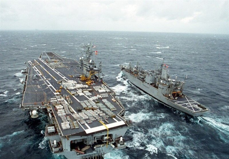Two naval vessels sail side by side in choppy open ocean waters under a cloudy sky, with cargo and equipment stacked across the aircraft carrier's flight deck.