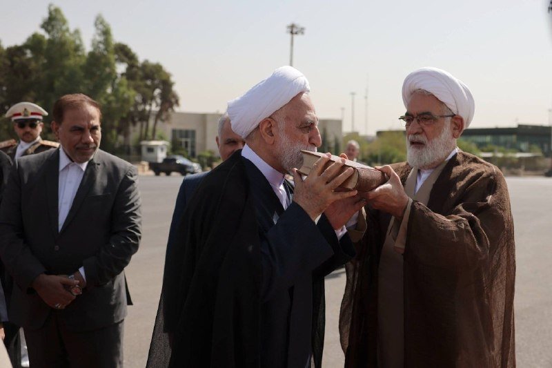 Two men wearing white turbans and robes stand outdoors on a paved area as one drinks from a small vessel held by the other.