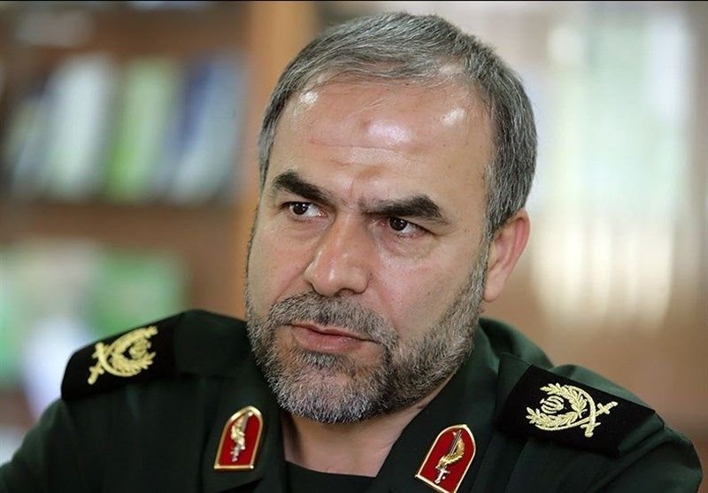 A bearded man with gray hair wears a green military uniform adorned with insignia on the collar and shoulders.