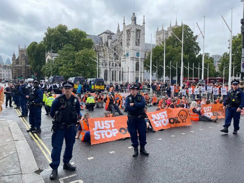 Protesters in orange vests sit on a street holding "JUST STOP OIL" banners while uniformed police officers stand guard on a road in front of a large Gothic-style building.