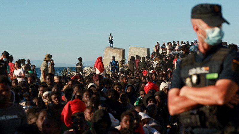 A large crowd of people gathers along a rocky coastline as a masked law enforcement officer in uniform stands watch in the foreground.