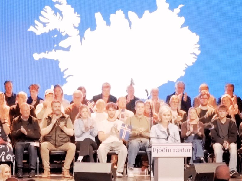 A group of people sit on stage in front of a large screen displaying the white outline map of Iceland, with some holding Icelandic flags and a podium reading "Þjóðin ræður."